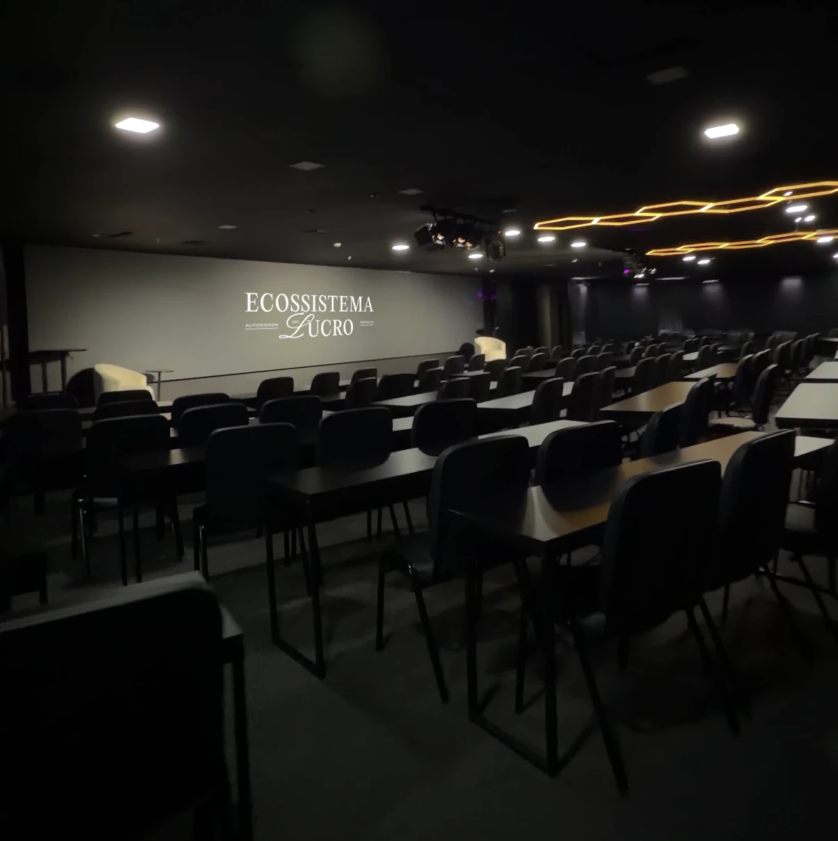 A sala da imersão: mesas e cadeiras voltadas para o telão com o logo do Ecossistema do Lucro